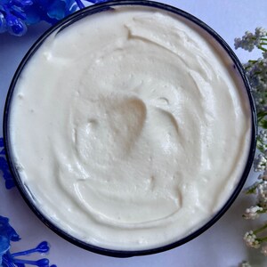 May include: A close-up of a white, creamy body butter in a blue jar. The butter has a smooth, whipped texture.