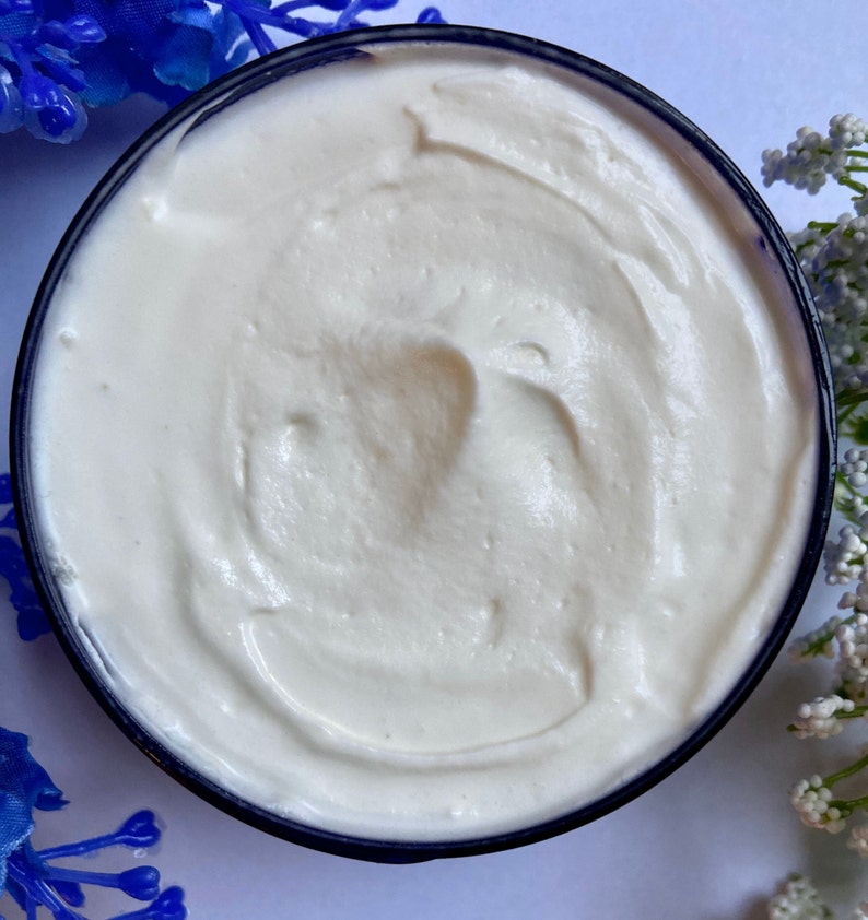 May include: A close-up of a white, creamy body butter in a blue jar. The butter has a smooth, whipped texture.