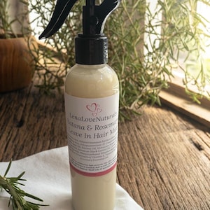 May include: A clear bottle of LenaLoveNaturals Batana & Rosemary Leave In Hair Mist with a black spray nozzle. The label includes ingredients and product information. The bottle sits on a white cloth, with rosemary sprigs in the background.