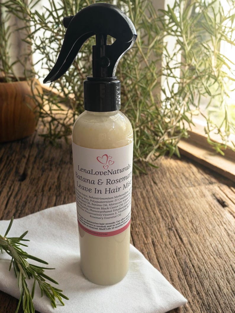 May include: A clear bottle of LenaLoveNaturals Batana & Rosemary Leave In Hair Mist with a black spray nozzle. The label includes ingredients and product information. The bottle sits on a white cloth, with rosemary sprigs in the background.
