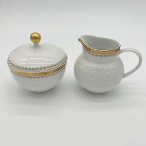 May include: A white porcelain sugar bowl with a lid featuring a gold sphere finial and a matching creamer pitcher. Both pieces are decorated with a gold band and a subtle embossed circular pattern around the exterior of the white ceramic body.