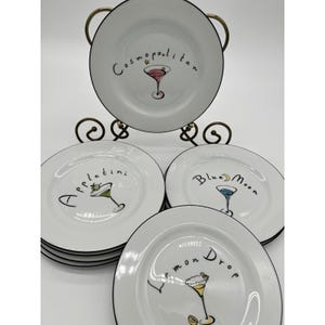 May include: A set of four white ceramic plates with black rims, each featuring a hand-drawn cocktail illustration and text. The designs include a pink Cosmopolitan, a green Appletini, a blue Blue Moon, and a yellow Lemon Drop cocktail.
