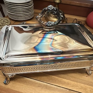 May include: A rectangular silver-plated chafing dish with a polished finish and a reflective surface. It features a decorative ornate handle on the lid, a pierced lattice-style gallery border, and four small, curved feet at the base.