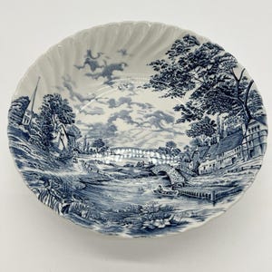 May include: A white ceramic bowl with a scalloped edge featuring a blue transferware pattern of a rural landscape. The scene depicts a river, a stone bridge, a small boat, houses, a church steeple, and trees under a cloudy sky in a classic style.
