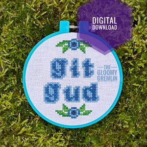 Peut inclure: Un cerceau de broderie bleu et blanc avec les mots "Sit  -The- Gloomy Gremlin  Gud" brodés en fil bleu. Le cerceau est entouré de mousse verte.