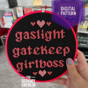 Peut inclure: Un cerceau de broderie noir avec une bordure rouge, présentant le texte "gaslight gatekeep girlboss" en fil rose. Le texte est entouré de petits cœurs roses.