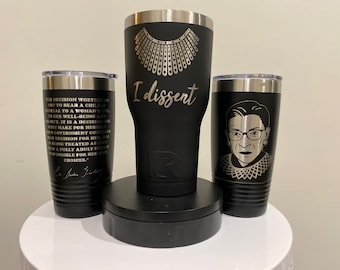 May include: Three black stainless steel tumblers with silver engravings. The center tumbler has the text "I dissent" and a design of a collar. The tumbler on the left has a quote from Ruth Bader Ginsburg. The tumbler on the right has a portrait of Ruth Bader Ginsburg.