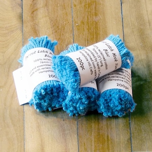 May include: Several bundles of vibrant blue latch hook yarn. Each bundle has a white label with text. The yarn is made from 100% reclaimed cotton and is 7.6 cm long. The bundles are arranged on a wooden surface.