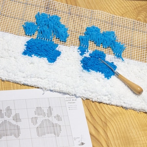 May include: A close-up of a white rug with two blue paw prints. The rug is being worked on with a latch hook tool. A printed pattern for the rug is visible below the rug.