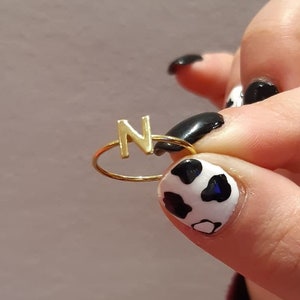 May include: A gold ring with the letter "N" on it. The ring is being held by a hand with black and white cow print nail polish.