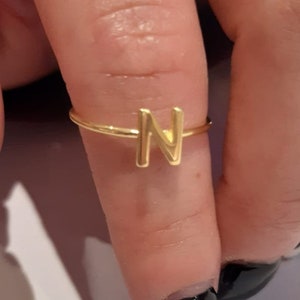 May include: A gold ring with the letter N on it. The ring is on a person's finger.