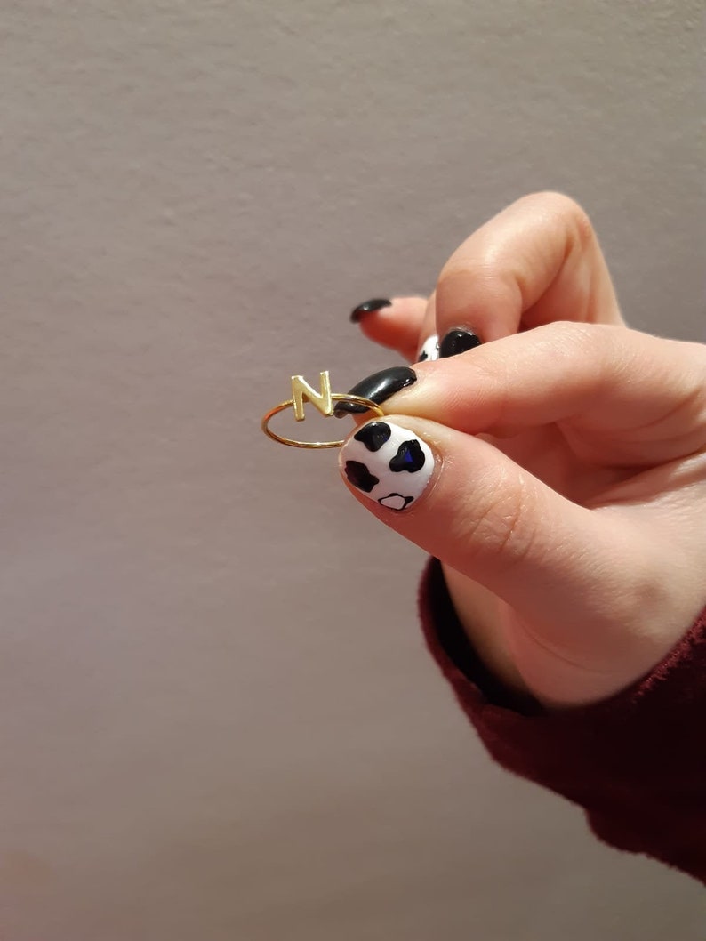 May include: A gold ring with the letter "N" on it. The ring is being held by a hand with black and white cow print nail polish.