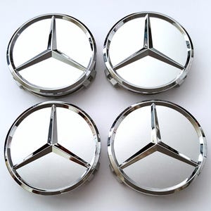 May include: Four chrome-finished Mercedes-Benz wheel center caps. Each cap features the iconic three-pointed star emblem in the center, surrounded by a polished silver ring. The caps are arranged in a square formation against a white background.