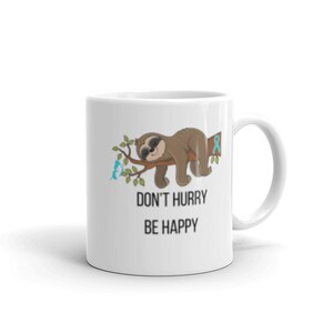 Könnte beinhalten: Weiße Keramiktasse mit einem schlafenden Faultier auf einem Ast mit blauen Schmetterlingen. Die Tasse trägt den Text "Eile nicht, sei glücklich".