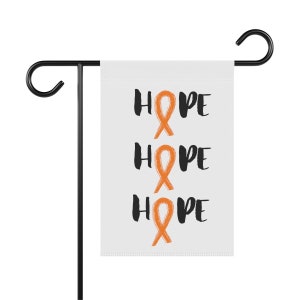 May include: A white garden flag with the word "HOPE" in black script, repeated three times with an orange ribbon graphic in place of the "O". The flag is displayed on a black metal garden stake.