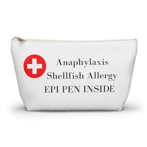 Può includere: Pochette bianca con cerniera con un simbolo a croce rossa e le parole "Anaphylaxis Shellfish Allergy EPI PEN INSIDE" in nero. La pochette ha una cerniera dorata ed è progettata per trasportare un EpiPen.