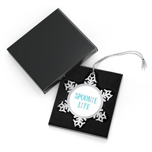 May include: A silver snowflake ornament with a blue and white circle in the center that says "Spoonie Life" with a spoon and fork graphic. The ornament is in a black box with a silver string.