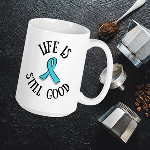 Könnte beinhalten: Weiße Keramik-Kaffeetasse mit einem türkisfarbenen Band und dem Text "Life is still good".