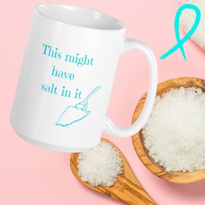 Könnte beinhalten: Eine weiße Keramiktasse mit einem blauen Design, auf dem "This might have salt in it" steht und eine Zeichnung eines Löffels, der Salz schöpft. Die Tasse steht auf einer rosa Oberfläche mit einer Schüssel Salz und einem Holzlöffel mit Salz.