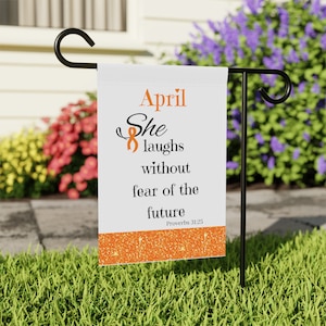 May include: A white garden flag with the text "April She laughs without fear of the future" in black and orange. The flag has an orange glitter border and is displayed on a black metal stand in a garden setting.