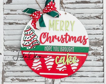 Pode incluir: Um letreiro de Natal redondo branco, vermelho e verde. Diz "Merry Christmas" e "Hope You Brought Cake". Uma fita vermelha e verde com bolinhas brancas é presa com um laço de barbante. Para decoração festiva.