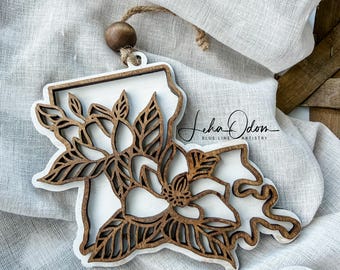 May include: A layered wooden ornament shaped like the state of Louisiana, featuring a white base and a dark brown laser-cut overlay of magnolia flowers and leaves. A small wooden bead is attached to the top with a jute twine hanging loop.