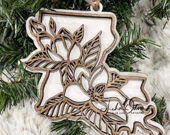 May include: Wooden Christmas ornament in the shape of Louisiana, featuring a magnolia flower design. The ornament is made of layered wood with a white and brown finish, and includes the text "Leha Odom BLUE LINE ARTISTRY". It hangs from a twine loop.