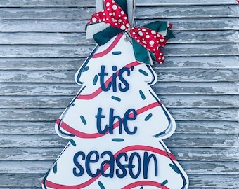Pode incluir: Uma placa de madeira em forma de árvore de Natal com o texto "'tis the season" em azul. A placa é branca com detalhes em vermelho e verde e um laço de bolinhas vermelho e branco. Uma corda de juta é anexada para pendurar.