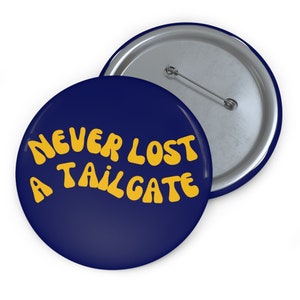 Puede incluir: Un botón azul con el texto "NEVER LOST A TAILGATE" en letras amarillas.
