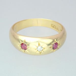 Può includere: Un anello d'oro con tre pietre preziose. Due gemme rosse affiancano una gemma bianca centrale, ciascuna incastonata in un design a stella. L'anello ha una fascia arrotondata e una finitura lucida.
