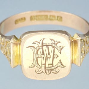 May include: A gold signet ring with an engraved monogram of the letters "W.E." on the top. The ring has a square-shaped top and a decorative band.