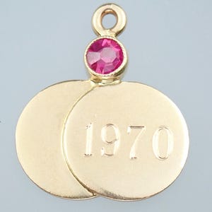 Puede incluir: Colgante dorado con una gema rosa brillante y dos discos dorados superpuestos. El año "1970" está grabado en uno de los discos. El colgante tiene un lazo para sujetarlo a una cadena.