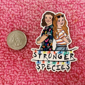 May include: A sticker featuring two cartoon women with their arms crossed. One wears a colorful shirt, the other a striped shirt and sunglasses. The text "STRONGER SPECIES" is below, with colorful lights.