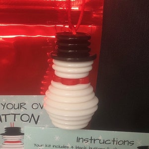 May include: A white snowman ornament made of stacked buttons with a red scarf and a black button hat. The ornament is hanging from a red string.