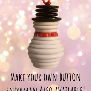 May include: A white button snowman ornament with a red hat and black button eyes. The snowman is hanging from a red string. The text "MAKE YOUR OWN BUTTON SNOWMAN ALSO AVAILABLE!" is at the bottom of the image.