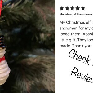 May include: A white snowman ornament made of stacked buttons with a red ribbon around its neck. The ornament is hanging from a red string on a green Christmas tree. The text "Check out our Reviews!" is visible in the background.