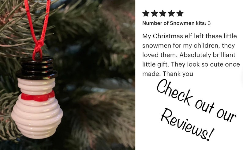 May include: A white snowman ornament made of stacked buttons with a red ribbon around its neck. The ornament is hanging from a red string on a green Christmas tree. The text "Check out our Reviews!" is visible in the background.