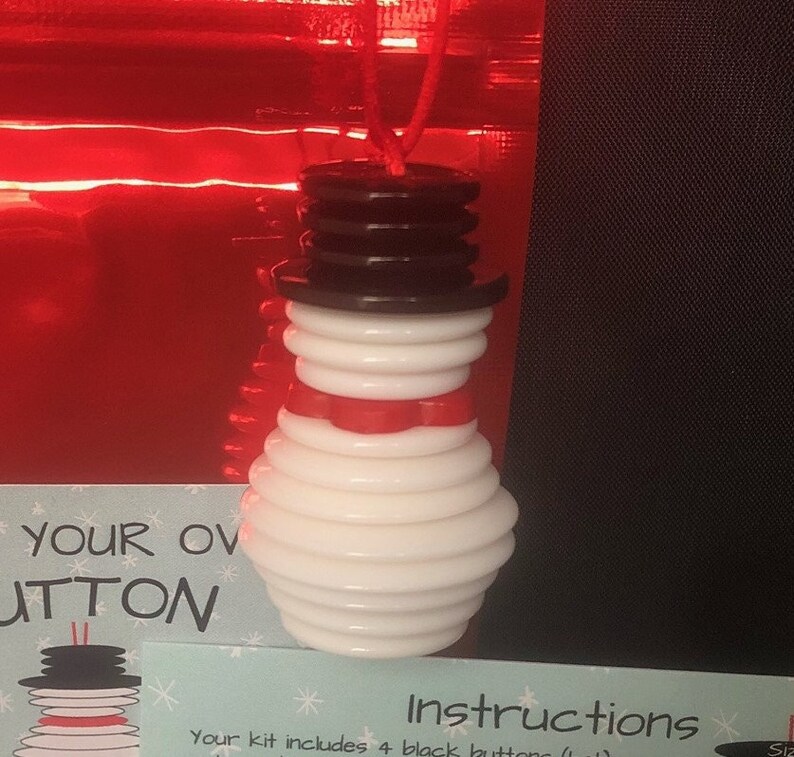 May include: A white snowman ornament made of stacked buttons with a red scarf and a black button hat. The ornament is hanging from a red string.