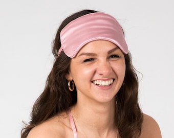 May include: A woman wearing a pink satin sleep mask, a pink satin top, and gold hoop earrings. She is smiling and looking at the camera.