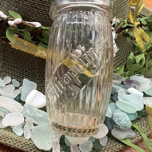 Op de afbeelding: Een heldere glazen pot met verticale ribbels, met een label met de tekst "Undum". De pot is omgeven door zee glas in verschillende tinten blauw, groen en wit. Een bloemenkrans en gouden lint zijn op de achtergrond.