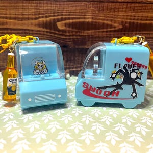 May include: Two light blue toy trucks with clear tops, each with a yellow chain and a miniature Corona beer bottle attached. One truck has a cartoon character, the other has the word "Swoon" and a graphic.
