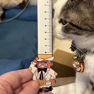 May include: A white and gray cat with a bell collar looks at a ruler with a cartoon character on it. The ruler is 8 cm long and has black markings. The cartoon character is wearing a white suit, a black bow tie, and a yellow hat. The character is holding a yellow star.