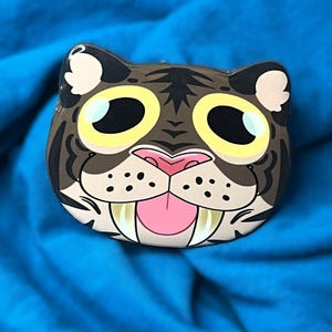 May include: A cartoon-style tiger face pin with a brown and tan color scheme. The pin features large yellow eyes, a pink tongue, and white fangs. The pin is set against a blue background.