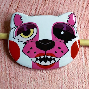 May include: A white, pink, and black cartoon cat face button with a menacing grin. The button features a white background with pink stripes and accents, red cheek circles, and a black nose.
