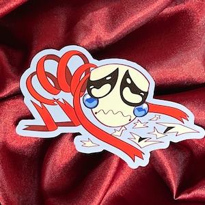 May include: A sticker featuring a cartoon character with a white face, large black eyes, and blue teardrops. Red ribbons and star-shaped elements surround the character. The sticker has a light blue outline and is set against a red background.