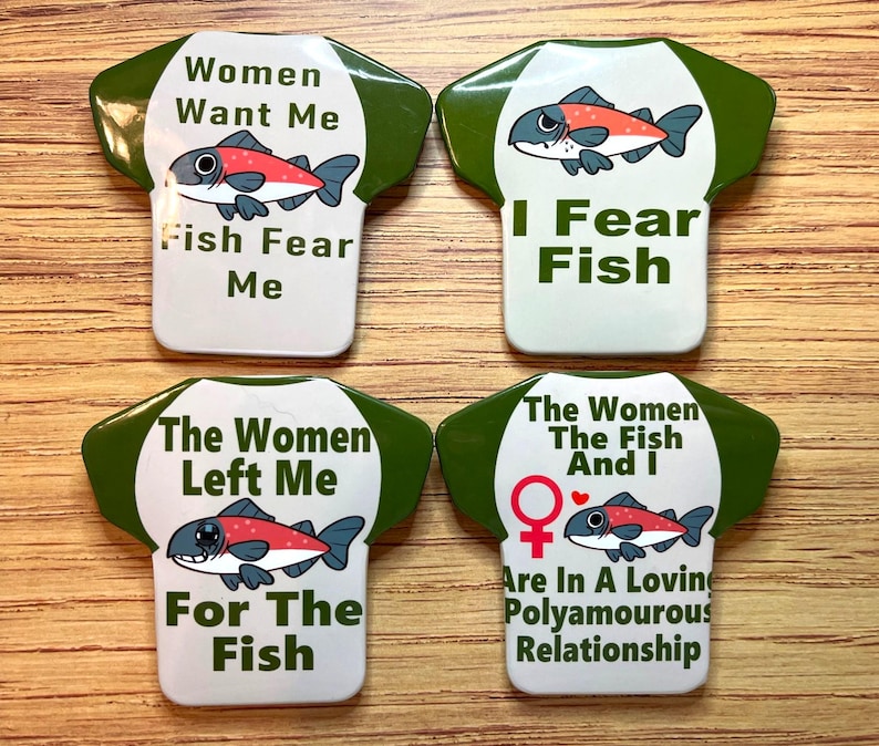 May include: Four white, shirt-shaped pins with green trim and various phrases and fish illustrations. The phrases include "Women Want Me, Fish Fear Me," "I Fear Fish," "The Women Left Me For The Fish," and "The Women, The Fish And I Are In A Loving Polyamorous Relationship."