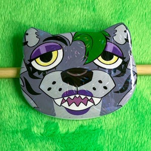 May include: A gray, circular pin featuring a stylized wolf face with yellow eyes, purple accents, and green hair. The wolf has a fierce expression with sharp teeth and a purple mouth. The pin is set against a green, textured background.