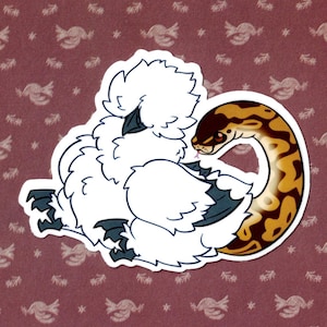 Op de afbeelding: Een wit, pluizig kippetje met zwarte ogen en een zwarte snavel is opgerold met een bruine en gele slang. De slang is om het lichaam van het kippetje gewikkeld en heeft een zwart oog.