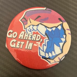 Puede incluir: Un botón redondo rojo con el texto blanco "Go Ahead, Get In". El botón presenta una ilustración de dibujos animados de una boca abierta con dientes azules y una cabeza morada y beige.