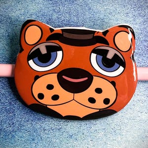 May include: A cartoon-style, orange and brown bear face with blue eyes, black accents, and a pink nose. The bear has a black hat and is set against a blue background. The image is a close-up of a decorative item.